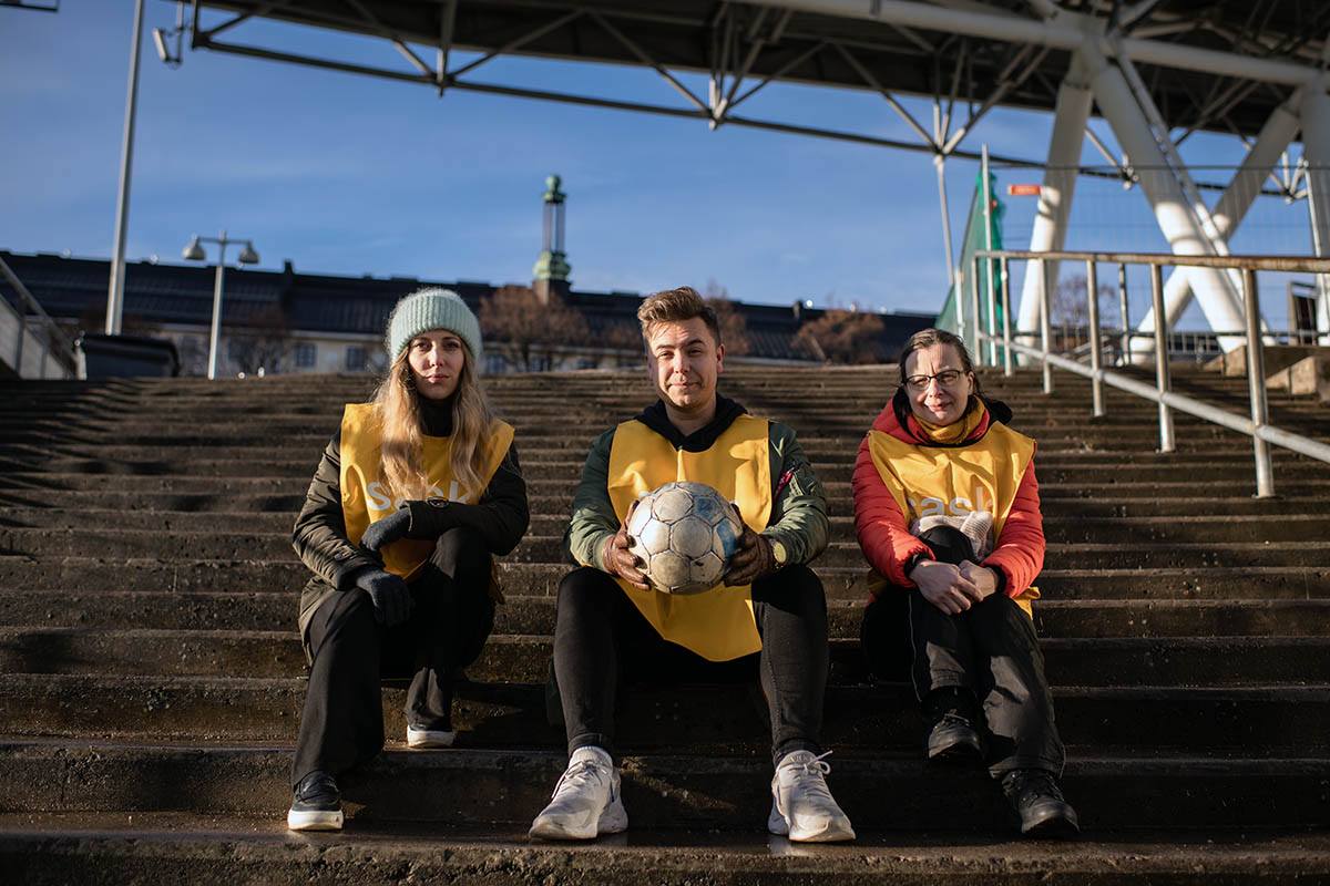 SASKin vapaaehtoistoimijat lVanessa Westerlund, Uula Neitola ja Katri Korhonen istumassa urheilustadionin portailla keltaiset SASK-liivit päällään.