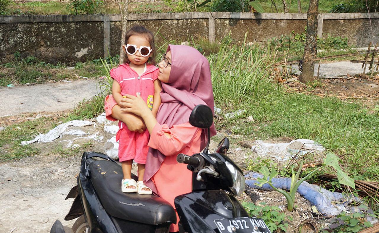 Indonesialainen nainen käsivarret kietoutuneena mopon päällä seisovan lapsensa ympärille. Molemmilla on vaaleanpunaiset vaatteet- Äiti katsoo lasta, lapsi katsoo kameraan.